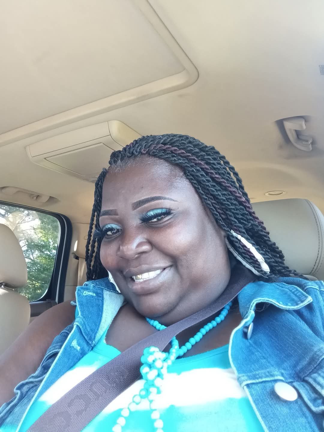 Woman smiling inside a car, wearing blue and turquoise clothing with beaded necklace and braided hair with turquoise accents