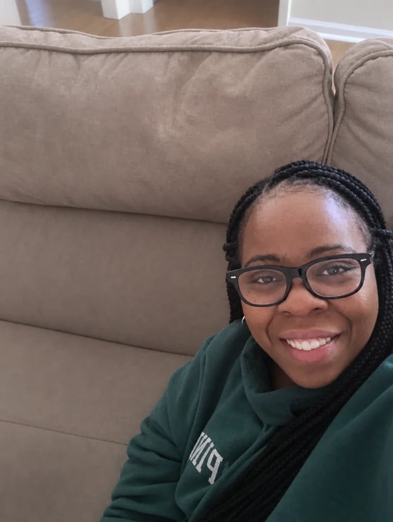 Smiling young woman wearing glasses and a green jacket, sitting on a beige couch indoors