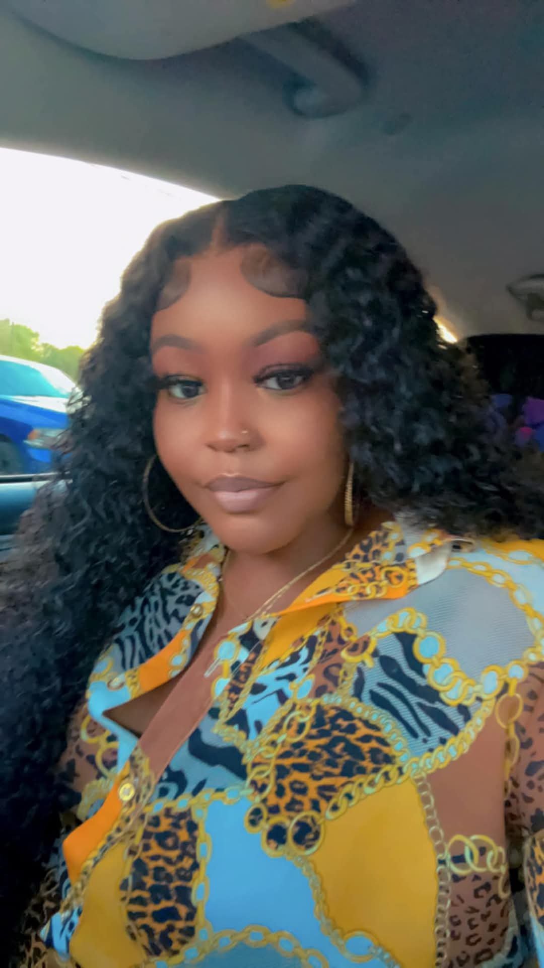 Woman with long black wavy hair wearing gold hoop earrings and a colorful patterned shirt, taking a selfie indoors