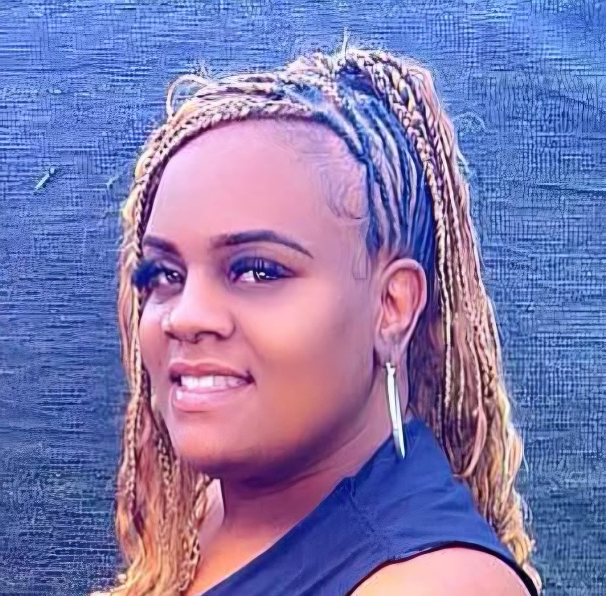 Portrait of a smiling woman with braided hair and hoop earrings wearing a blue sleeveless top against a textured blue background
