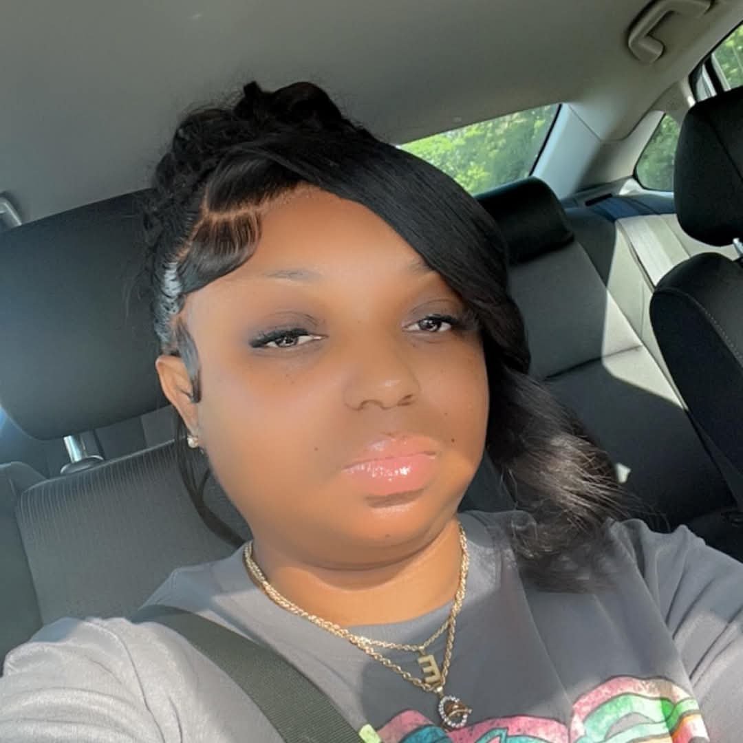 Woman with black wavy hair and gold necklace sitting in a car, wearing a gray shirt and looking at the camera